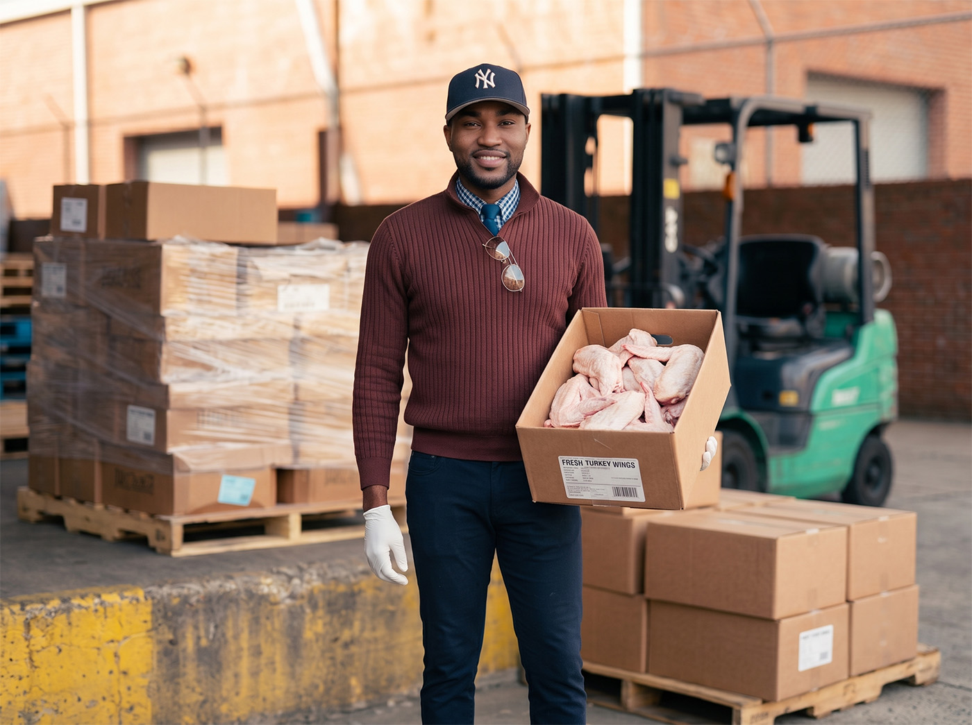 Man at a loading dock holding a box of fresh turkey wings, with a forklift and pallets in the background